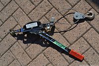 Side view of a manual Come-A-Long winch showing the red plastic handle grip, steel cable, and hooks.