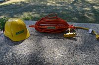 Three items displayed on concrete surface outside: a yellow hard hat, a coiled orange extension cord with multi-socket plug, and a yellow 4-outlet multi-plug power strip.