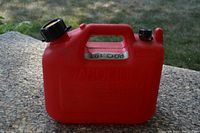Red gasoline jerry can with black screw-on top cap, integrated handle, and vent cap seen from the front/side angle on a stone surface outdoors.