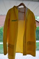 Front view of yellow poly vinyl rain jacket with hood, hanging on a hanger outdoors.