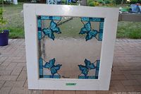 Square stained glass window featuring five blue butterflies with blue and clear textured glass, framed in white wood.