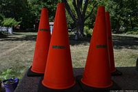 Four orange traffic cones arranged outdoors on a flat surface, each with a black base and markings 'LOT 001'.