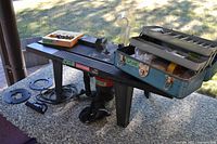 Front angled view of black Craftsman router table on legs with mounted router visible beneath, showing blue metal toolbox, wooden cases with router bits, plastic guard and various accessories on the table surface.
