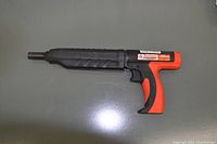 Side profile view of Ramset Mastershot tool showing black and orange pistol grip with a long black barrel, on a flat surface.