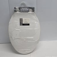 White plastic toilet seat wrapped in clear plastic packaging with 'Plastic Plastique' label visible at hinge.