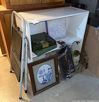 Small white bookshelf with folding black step stool leaning on it, assorted boxes and framed picture on floor.