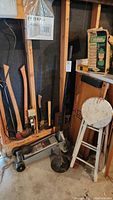Wide view of garage corner showing axes and hatchets on wooden rack, hydraulic floor jack on floor, auto creeper, and wood folding stool.