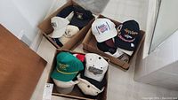 Three boxes filled with assorted ball caps of various sports teams and events, showing a diverse collection.