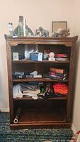 Front view of a dark brown wooden 4-shelf bookcase partially loaded with books, fabric, and decorative items on top.