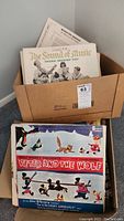 Cardboard box filled with vintage vinyl LPs including 'The Sound of Music' original Broadway cast album and Disney 'Peter and the Wolf' album in front with illustrated cover showing characters.