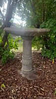Full side view of the concrete bird bath standing on a fluted column base among garden mulch and greenery.