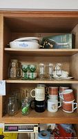 Wide view of wooden cabinet shelves showing Corningware dish, green goblets, clear glasses, mugs with patterned designs, and more glassware
