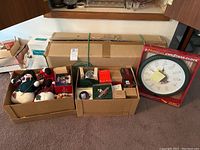Wide view of three boxes on floor showing antique ornaments, bubble lights, an oversized boxed musical Christmas clock, and holiday decor items