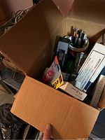 Open cardboard box containing assorted office supplies including pens, markers, tape dispenser, and Kodak projector slide boxes