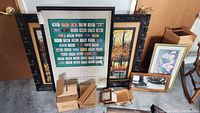 View of lot showing multiple framed artworks, a Smithsonian poster, small mirrors with boxes, and various framed paintings leaning against a wall.
