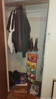 Closet view showing cowboy boots on a box, hangers with various jackets and sweaters, Peanuts hanging shoe organizer, and boxes of sports apparel beneath.