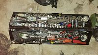 Top view of open metal tool box showing organized socket wrench sets, screwdrivers, and hand tools