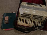 Accordion and case open showing keys, bass buttons and black leather straps alongside books and metronome