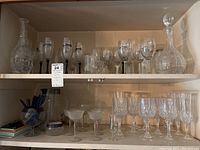 Wide shelf view displaying a large etched decanter on the right, multiple styles of etched and cut crystal wine and cocktail glasses arranged on two shelves, and a glass holding several glass swizzle sticks on the lower left.