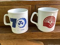 Two white ceramic mugs side by side. Left mug has blue '70' with trefoil emblem and dates 1910-1980. Right mug with red United Way logo.