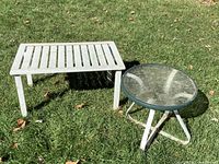 Two metal garden patio tables: one white metal rectangular table with slatted top and one round metal table with glass top and triangular legs, placed on grass outdoors.