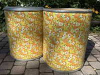Two vintage-style metal storage canisters with yellow, orange, white floral pattern on the outside, placed on paved ground with greenery in background.