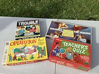 Photo of 4 classic board games in their boxes: Trouble, Scrabble for Juniors, Operation, and Teacher's Quiz arranged on a table outdoors