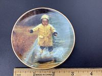 Front of miniature collector plate showing child in yellow raincoat splashing in puddle, signed Donald Zolan