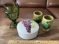 Set of one olive green pressed glass goblet, two vintage green ceramic vases, and one round ceramic lidded container arranged with ruler for scale.