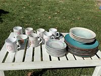 Photo of assorted mugs, plates, and bowls displayed on white slatted table outdoors on grass showing patterns and colors.