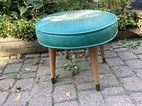 Full view of the round foot stool showing turquoise vinyl top with peeling damage and four wooden legs with darkened tips and metal ferrules.