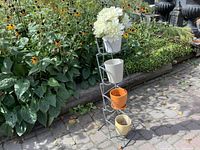 Metal plant stand with planting pots placed inside some holders, standing on a paved outdoor surface next to green plants.