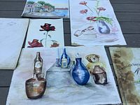 A collection of watercolour paintings displayed on a gray surface, showing bottles, vases, a teddy bear, flowers, and a coastal scene.