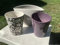 Photo showing all three ceramic pots on a white surface outdoors in sunlight: a white elongated boat-shaped pot, a white pot with floral pattern, and a purple pot with circular holes near top.