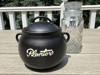 Two vintage Planters Peanuts jars including a black ceramic jar and a vintage clear glass canister with lid.