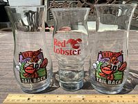 Three Red Lobster beer glasses on a table with a ruler for scale. Two glasses have a colorful cartoon lobster holding a beer mug with 'Red Lobster Tail Back' text. The middle glass has a large red lobster logo and 'Red Lobster' text in red.