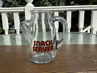 Side view of clear glass pitcher with handle and red "SNACK SERVER" lettering on side, placed on wooden table outdoors.