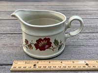 Off-white ceramic creamer pitcher with red and brown floral design shown from side on wooden surface with ruler for scale approximately 4 inches tall.