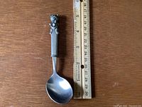 Full view of Royal Selangor Teddy Bear Picnic baby spoon laid next to a measuring stick for scale, showing spoon length about 5.5 inches.