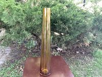 Tall amber glass vase placed outdoors on a brown surface against green shrubbery background, showing the full height and color of the vase.