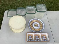 Photo showing 5 Tupperware containers of various sizes with green seals, 2 decorative serving trays with artwork, one round divided serving tray with blue and brown pattern, and a white Lazy Susan stand.