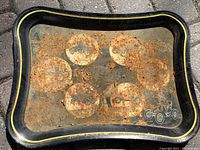Top view of vintage 1960s black metal serving tray with scalloped edges and yellow border, surface heavily rusted obscuring some antique car drawings.