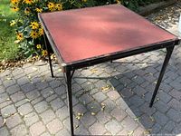 Square vintage card table outdoors on paved surface, showing reddish-brown tabletop and metal folding legs with rust.