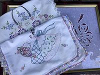 Close-up of two embroidered linen table runners showing detailed floral and lady motifs with crocheted trim.