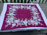 Maroon linen tablecloth with white floral rose and leaf pattern laid flat on a wooden deck surface, showing full design and white border.