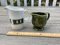 Two vintage creamers side by side with a ruler for size reference, showing green ceramic Japanese creamer and white Johnson Bros. creamer with olive green pattern.
