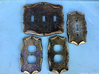 Four vintage metal light switch cover plates displayed on a light blue surface with two outlet covers, one single toggle cover, and one triple toggle cover.