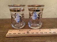 Pair of vintage hand-painted shot glasses with blue flower designs and gold band accents shown side by side with a ruler for size reference