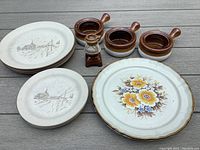 Photo of vintage stoneware dishes showing several plates with Manoir scenic design, floral design plates, three small handled brown stoneware bowls, and a Royal Sealy Spring branded salt or spice grinder on a wooden surface.