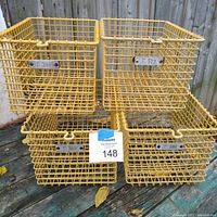 Four yellow coated wire locker baskets stacked two by two on a wooden surface outdoors, showing wear and metal number tags.